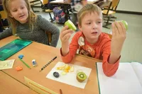 Zwei Kinder sitzen an einem Tisch in einem Klassenzimmer. Ein Junge hält einen grünen Apfel. Ein Mädchen beobachtet ihn. Verschiedene Früchte sind auf dem Tisch.