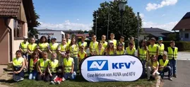 Eine Gruppe von Kindern und Erwachsenen in gelben Warnwesten posiert für ein Foto mit einem Banner, das 'AV KFV' vor einem Gebäude und Häusern zeigt.