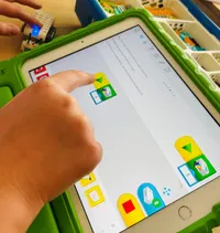 Eine Hand drückt auf einem Tablet mit grüner Abdeckung einen Knopf. Der Bildschirm zeigt verschiedene Symbole, darunter ein Häkchen, ein Ausrufezeichen und ein Dreieck.