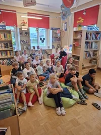 Eine Gruppe von Kindern sitzt auf Beanbags in einer Bibliothek, umgeben von Bücherregalen voller Bücher. Einige tragen Turnschuhe, während andere Sandalen tragen.