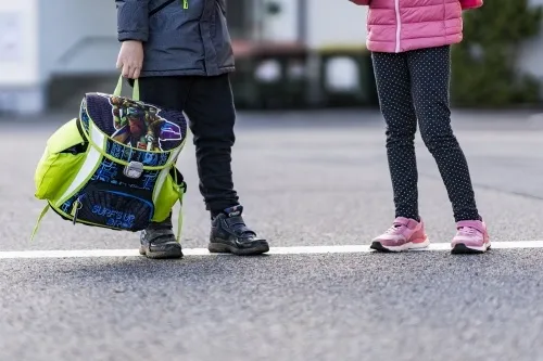 Zwei Kinder stehen auf der Straße, eines hält einen Rucksack mit einer Figur aus den Teenage Mutant Ninja Turtles. Sie tragen beide Turnschuhe und schwarze Hosen.