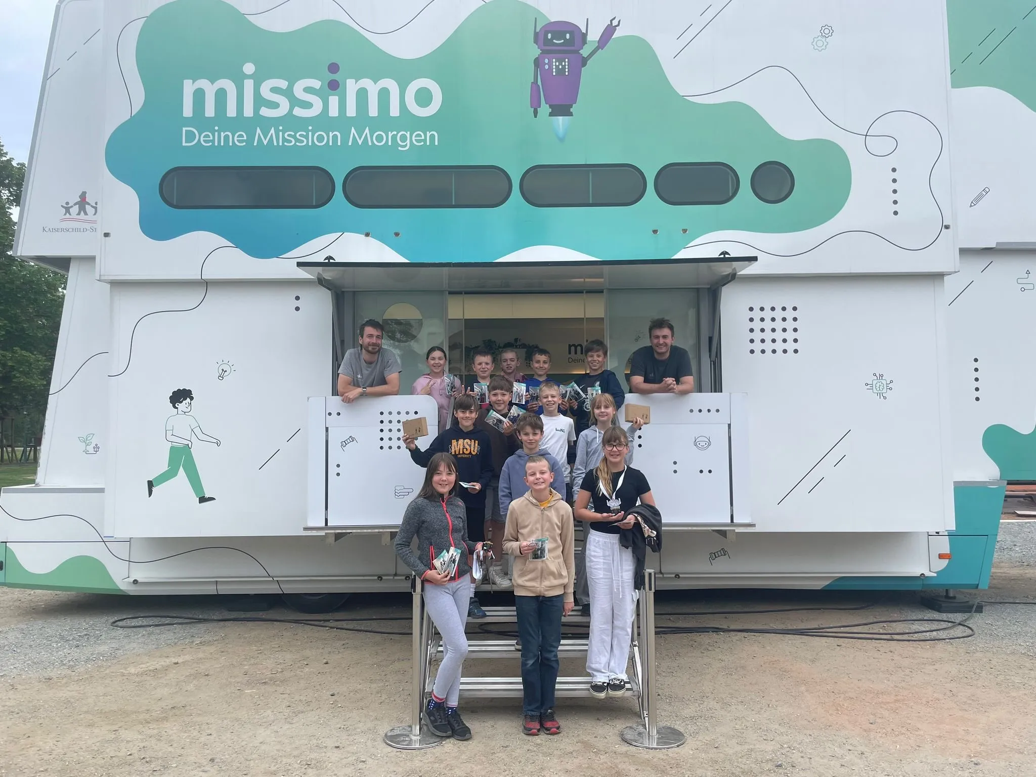 Eine Gruppe von Kindern und Erwachsenen steht vor einer mobilen Lerneinheit mit der Aufschrift Missimo. Sie alle lächeln und halten Bücher und Auszeichnungen in den Händen.