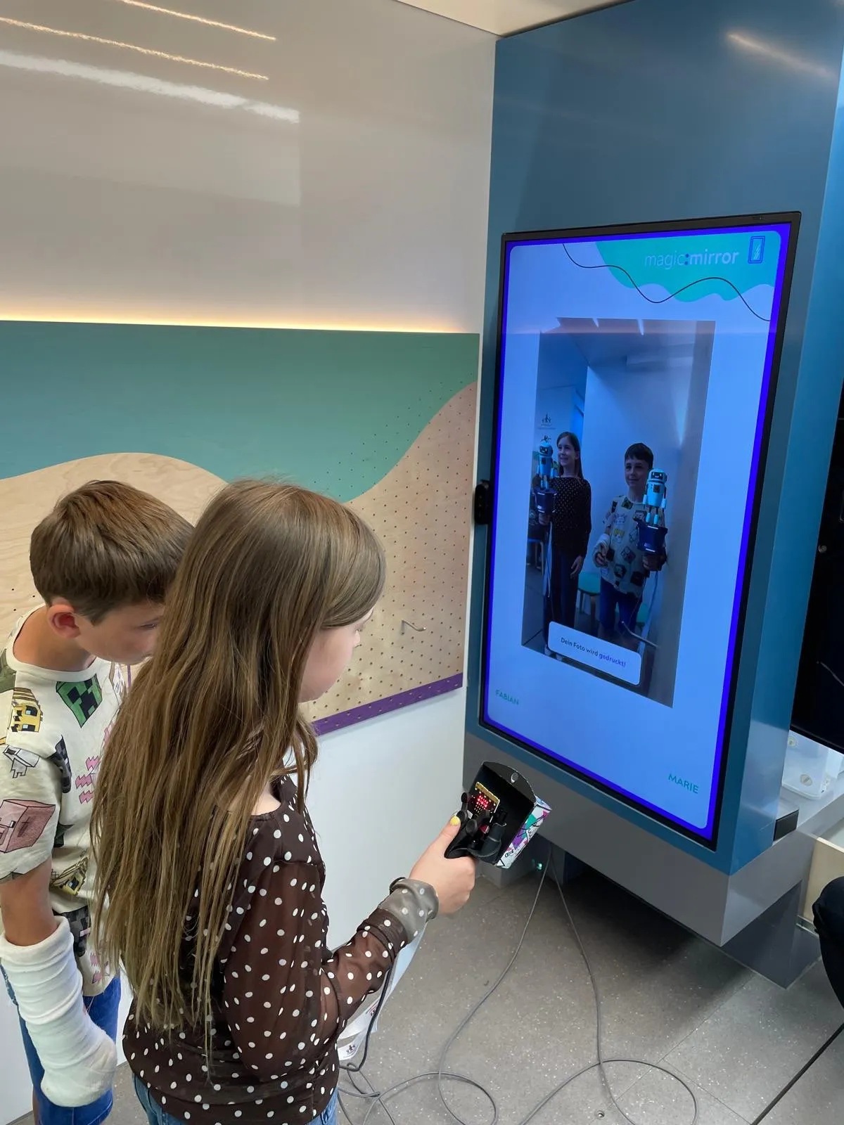Zwei Kinder benutzen einen Zauberspiegel, ein Mädchen hält eine Fernbedienung und ein Junge schaut auf den Bildschirm, der zwei Kinder mit Roboterspielzeug zeigt.