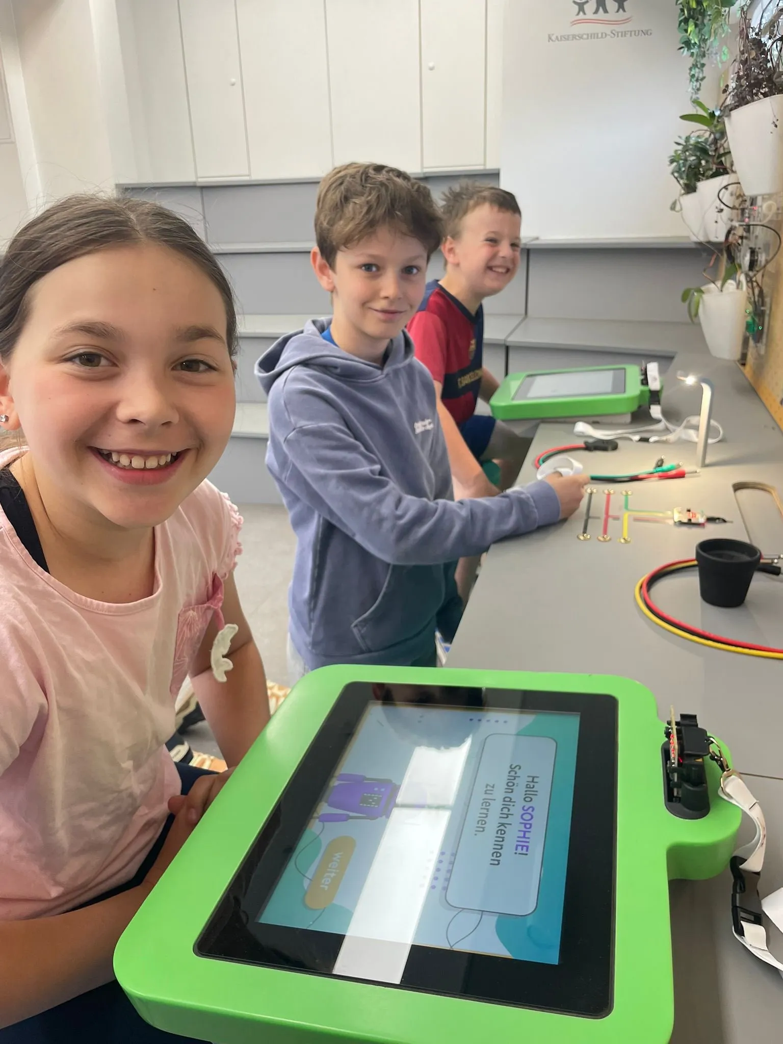 Drei Kinder lächeln und arbeiten an einem Projekt an einem Tisch mit einem grünen Tablet und Kabeln. Das Mädchen in Pink hält das Tablet.