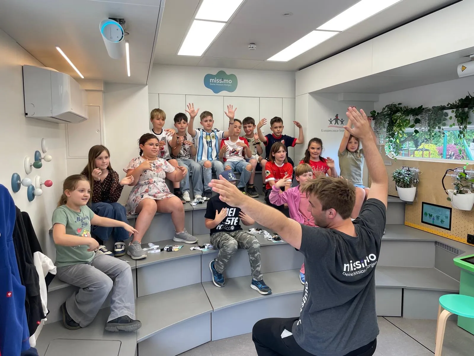 Eine Gruppe von Kindern und einem Mann macht eine Handübung in einem Raum. Einige sitzen auf einer Stufe, andere stehen. Ein Logo ist an der Wand.