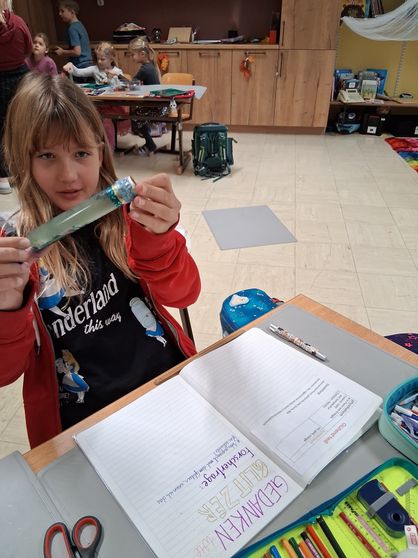 Ein junges Mädchen mit einer roten Jacke hält ein verpacktes Objekt in den Händen und sitzt an einem Schreibtisch mit Notizbuch und Stift in einer Klassenzimmerumgebung.