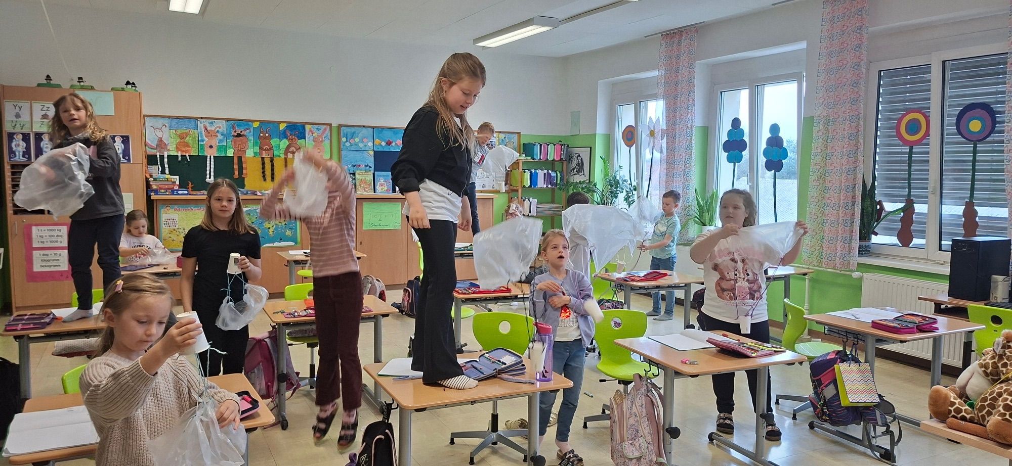In einem Klassenzimmer stehen Kinder auf Tischen. Ein Mädchen steht auf einem Tisch und hält ein weißes Objekt. Andere Kinder stehen auch auf Tischen und halten ähnliche Objekte. Ein Junge steht im Hintergrund, in der Nähe eines Regals mit bunten Flaschen.