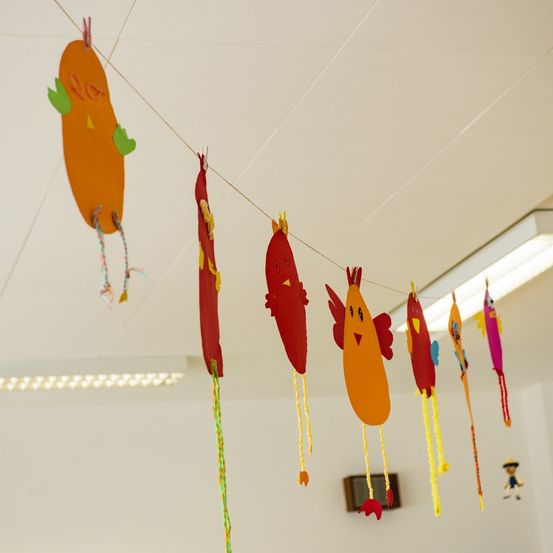 Eine Reihe bunter Papiervögel hängt von einer Decke mit weißen Fliesen, beleuchtet von einer Leuchtstofflampe rechts.