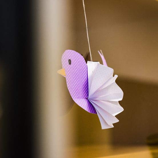Ein Papiervogel mit einem lila Körper und weißen Flügeln hängt an einer weißen Schnur und spiegelt sich in einem Fenster wider.