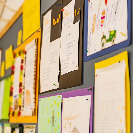 Ein Klassenbulletinbrett mit verschiedenen farbigen Papieren und Zeichnungen, darunter eine farbenfrohe Traktorzeichnung, die angeheftet sind.