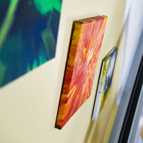 Ein leuchtendes orangefarbenes und gelbes abstraktes Gemälde hängt an einer cremefarbenen Wand, neben einem grün-blauen Gemälde.