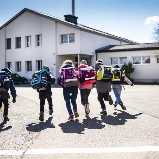 Mehrere Kinder mit Rucksäcken laufen auf ein weißes Gebäude mit vielen Fenstern zu. Ein Auto ist vor dem Gebäude geparkt.