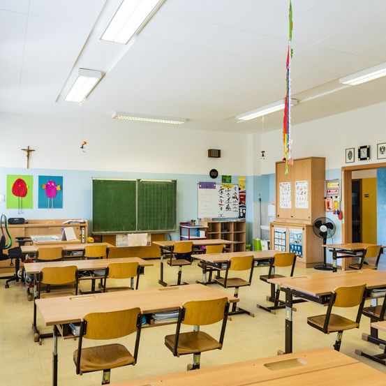 Ein leerer Klassenraum mit in Reihen angeordneten Tischen und Stühlen. Eine Tafel und eine Uhr sind an der Wand. Kunstposter und ein Kreuz schmücken die Wand. Lampen und ein Ventilator sind an der Decke.