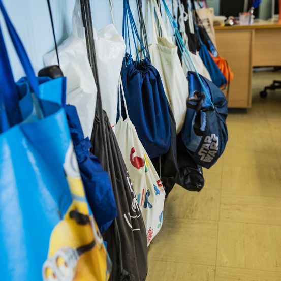 Eine Reihe von verschiedenen Taschen hängt an einer Wand, darunter einige mit bunten Designs, eine mit einem Star Wars-Logo und andere mit verschiedenen Logos. Ein Holzschreibtisch ist im Hintergrund zu sehen.