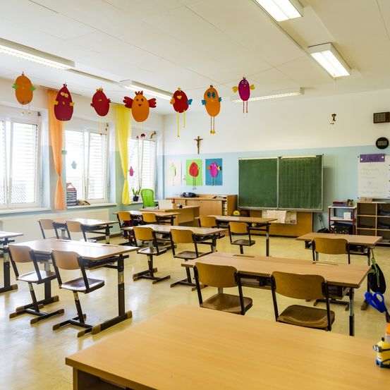 Ein leerer Klassenraum mit Holztischen und Stühlen, einer grünen Tafel und bunten Vogeldekorationen an der Decke. Der Raum hat Fenster mit orangefarbenen Vorhängen und eine weiße Decke mit Leuchtstofflampen.