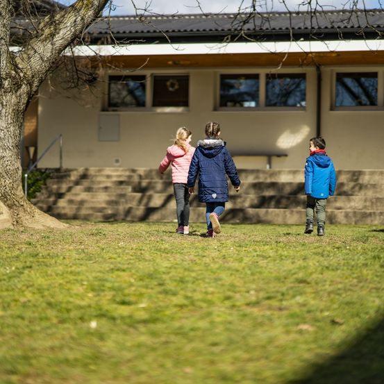 Drei Kinder in Wintermänteln gehen auf einem Grasrasen vor einem Gebäude mit Glasfenstern und einer Treppe.