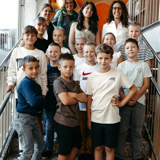 Eine Gruppe von Kindern und Erwachsenen posiert für ein Foto auf einer Treppe mit Metallgeländern. Sie alle lächeln und scheinen glücklich zu sein.