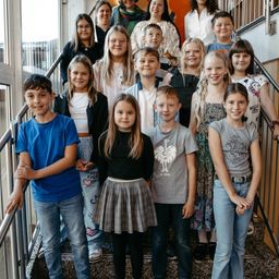 Eine Gruppe von Kindern und Erwachsenen steht auf der Treppe und lächelt für ein Gruppenfoto. Sie tragen Freizeitkleidung. Der Junge auf der linken Seite trägt ein blaues T-Shirt, während das Mädchen in der Mitte ein schwarzes T-Shirt und einen Rock trägt.