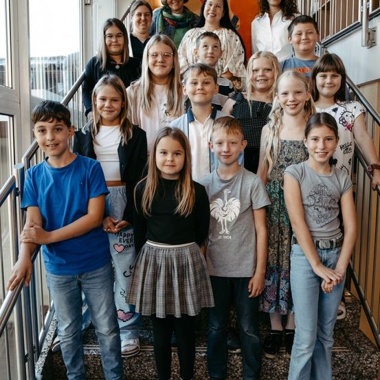 Eine Gruppe von Kindern und Erwachsenen steht auf der Treppe und lächelt für ein Gruppenfoto. Sie tragen Freizeitkleidung. Der Junge auf der linken Seite trägt ein blaues T-Shirt, während das Mädchen in der Mitte ein schwarzes T-Shirt und einen Rock trägt.