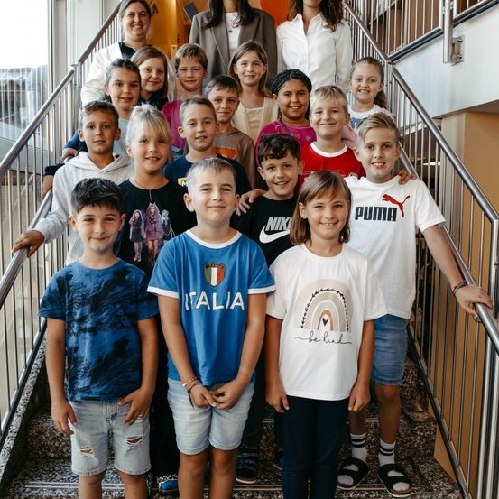 Eine Gruppe von Kindern mit zwei Erwachsenen lächelt auf einer Treppe. Einige tragen Markenshirts. Die Geländer sind aus Metall.