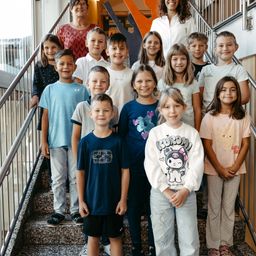 Eine Gruppe lächelnder Kinder und zwei Frauen stehen auf den Treppenstufen, wahrscheinlich in einer Schulumgebung, mit einem Baum und einem Gebäude im Hintergrund.