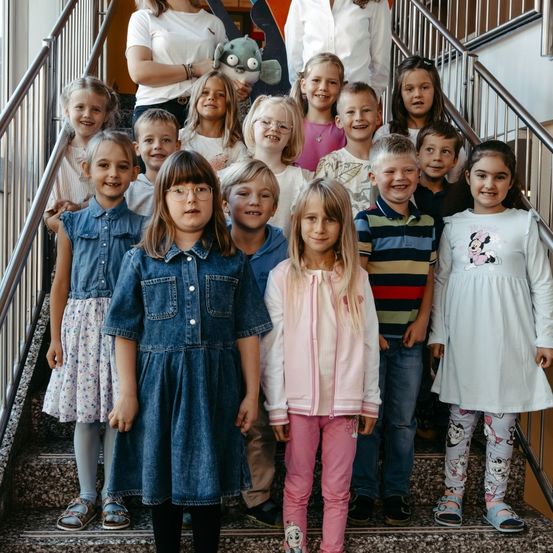 Eine Gruppe von Kindern und zwei Erwachsenen steht auf einer Treppe und lächelt für ein Foto. Ein Kind hält ein Kuscheltier.