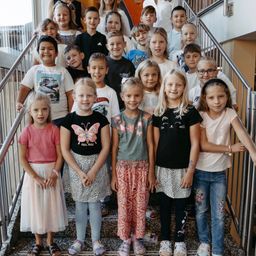 Eine Gruppe von Kindern steht auf einer Treppe und lächelt für ein Foto. Sie tragen verschiedene Kleidungsstücke, darunter T-Shirts, Röcke und Jeans. Die Treppe besteht aus Metall mit einem strukturierten Muster.