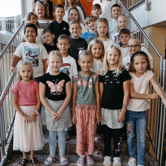 Eine Gruppe von Kindern steht auf einer Treppe und lächelt für ein Foto. Sie tragen verschiedene Kleidungsstücke, darunter T-Shirts, Röcke und Jeans. Die Treppe besteht aus Metall mit einem strukturierten Muster.