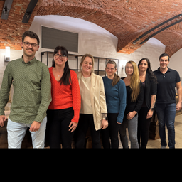 Eine Gruppe von sieben Menschen, darunter Männer und Frauen, steht in einem Raum mit Ziegelwänden und gewölbten Decken. Sie lächeln für ein Foto.