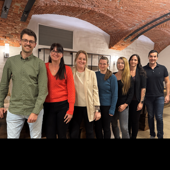 Eine Gruppe von sieben Menschen, darunter Männer und Frauen, steht in einem Raum mit Ziegelwänden und gewölbten Decken. Sie lächeln für ein Foto.