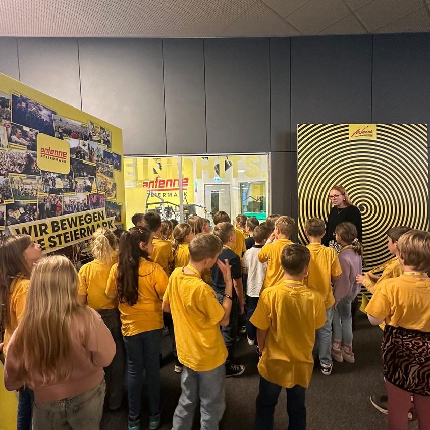 Eine Gruppe von Kindern in gelben Shirts und Turnschuhen ist um eine Frau versammelt, wahrscheinlich eine Lehrerin, in einem Raum mit gelben Wänden und einem Spiralmuster.