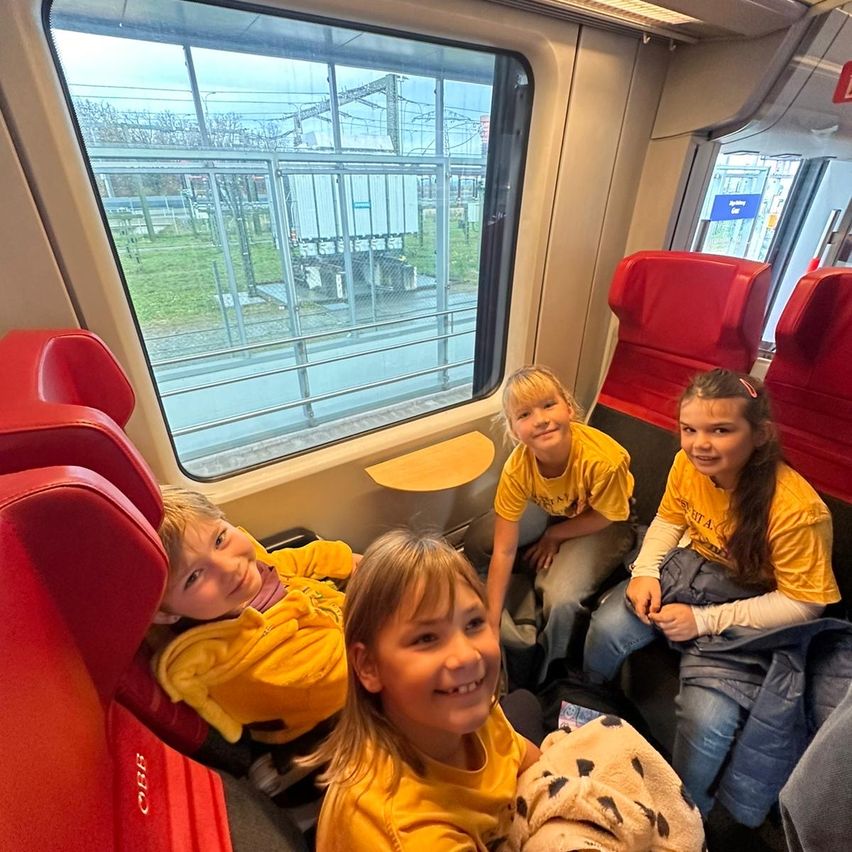 Vier Kinder in gelben T-Shirts sitzen in einem Zug. Ein Mädchen hält ein Kuscheltier. Der Innenraum des Zuges hat rote Sitze und ein Glasfenster mit Blick nach draußen.