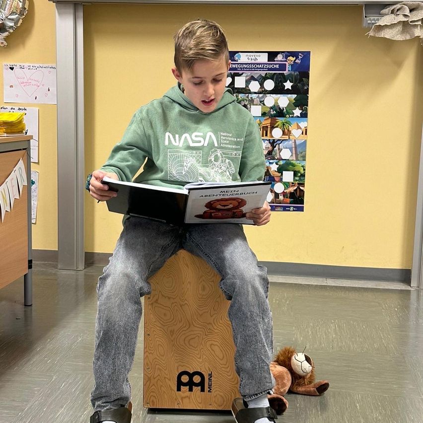 Ein Junge sitzt auf einem Holzhocker und liest in einem Buch mit dem Titel 'Mein Abenteuerbuch'. Er trägt eine NASA-Kapuzenjacke, mit einem Kuscheltier in der Nähe. Der Raum hat gelbe Wände und Bildungsplakate.