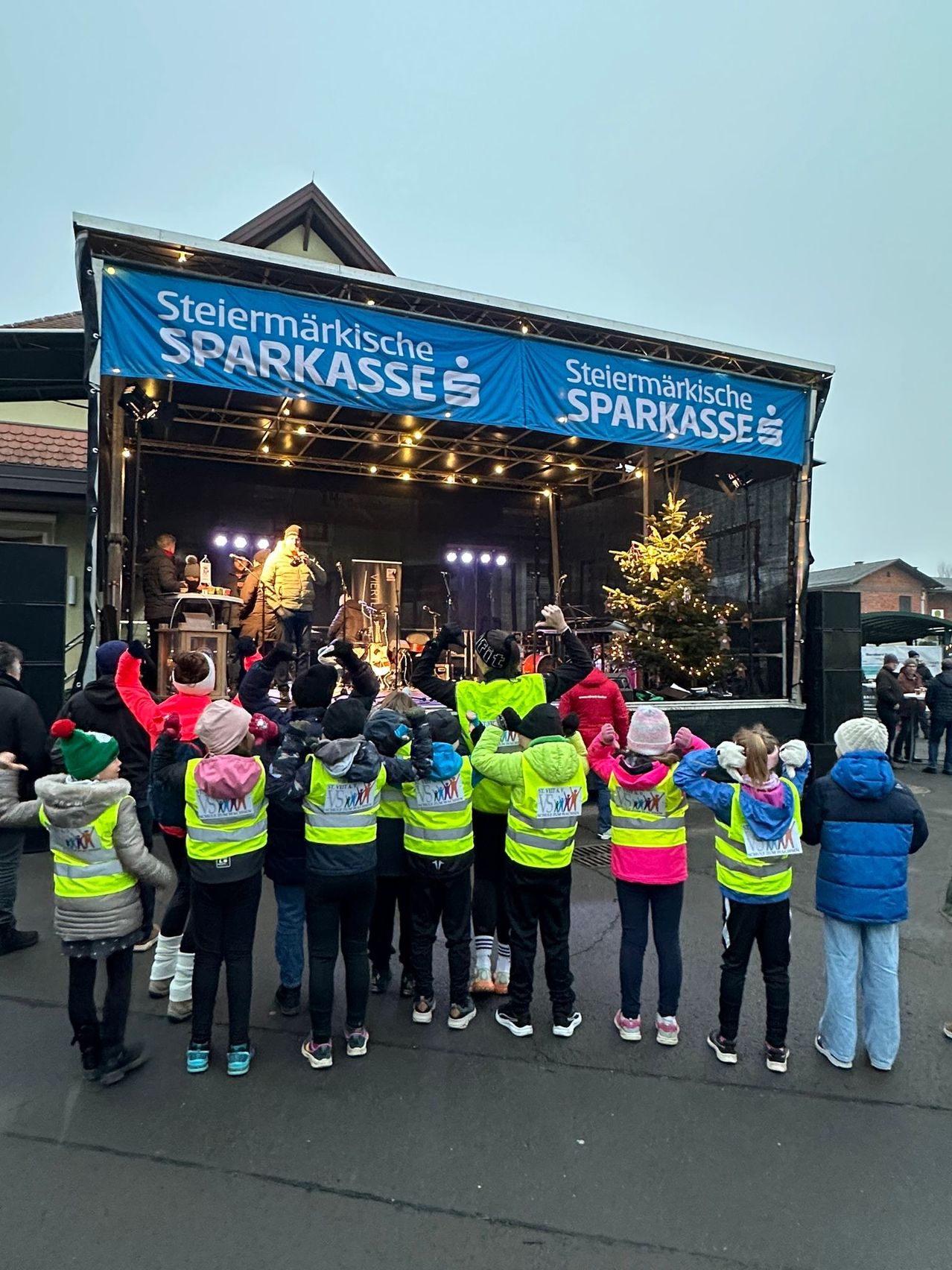 Eine Gruppe von Kindern in Sicherheitswesten versammelt sich vor einer Bühne mit einem Banner, auf dem 'Steiermarkische Sparkasse' steht. Dahinter steht eine Person mit Mikrofon auf der Bühne, und ein Weihnachtsbaum ist dekoriert.