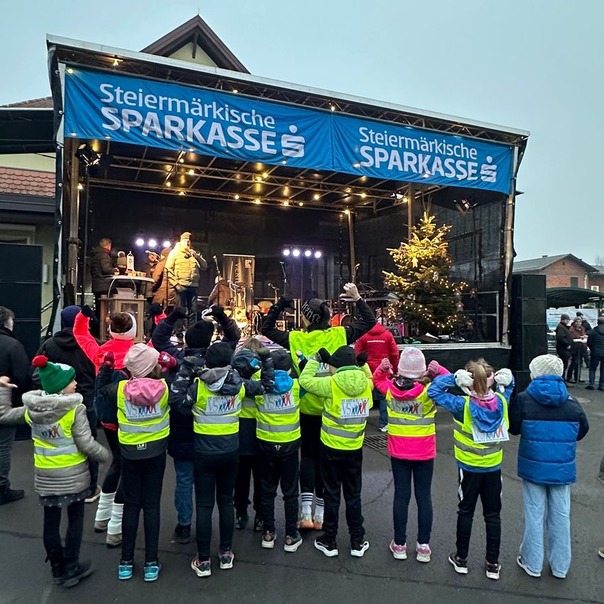 Eine Gruppe von Kindern in Sicherheitswesten versammelt sich vor einer Bühne mit einem Banner, auf dem 'Steiermarkische Sparkasse' steht. Dahinter steht eine Person mit Mikrofon auf der Bühne, und ein Weihnachtsbaum ist dekoriert.