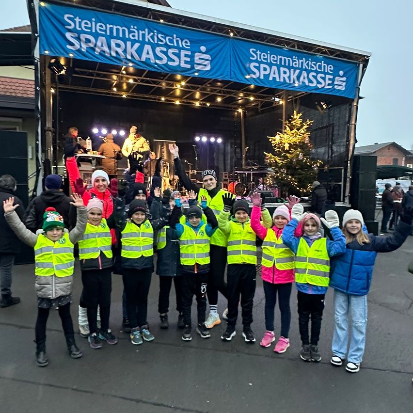 Eine Gruppe von Kindern in Warnwesten posiert für ein Foto vor einer Bühne mit einem Banner, das 'Steiermarkische Sparkasse' steht.