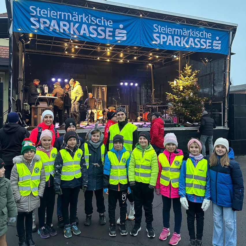 Eine Gruppe von Kindern in gelben Westen steht vor einer Bühne mit einem Banner, auf dem 'Steiermarkische Sparkasse' steht. Ein Weihnachtsbaum und ein Mikrofonständer sind auf der Bühne, mit Leuten auf der Bühne und weiteren Leuten, die hinter der Bühne stehen.
