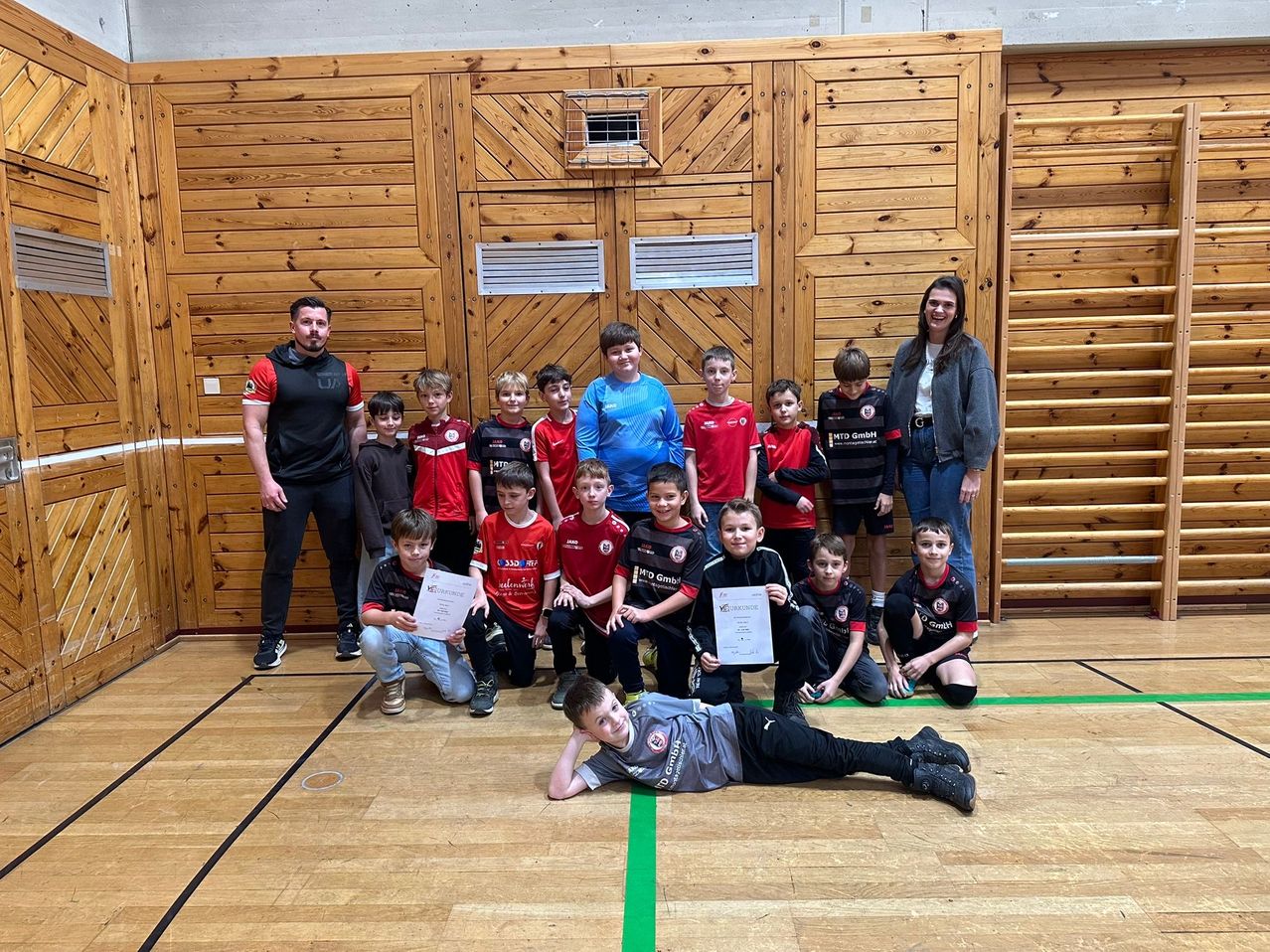 Eine Gruppe junger Jungen und Mädchen in Sportkleidung posiert für ein Foto mit ihrem Trainer, der in einer Turnhalle mit Holzwänden und einer grünen Linie auf dem Boden steht.