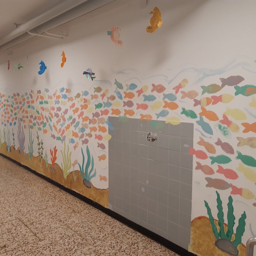 Ein Korridor mit einer farbenfrohen Unterwasserszene an der Wand, die verschiedene Fische, Seepferdchen und Korallenriffe zeigt. Die Wand hat in der Mitte einen gefliesten Bereich.