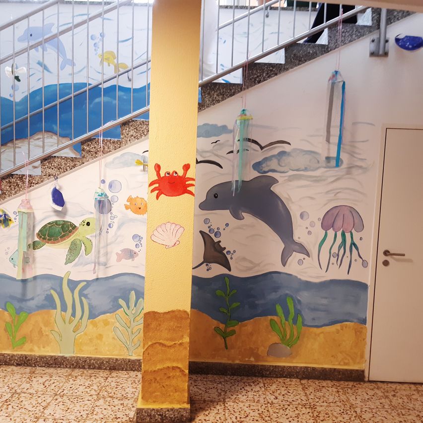 Eine Wand mit Meereslebewesen, darunter Delfine, Meeresschildkröten und Quallen, schmückt die Wände einer Treppe. Schnüre hängen von oben und verleihen einen verspielten Touch.
