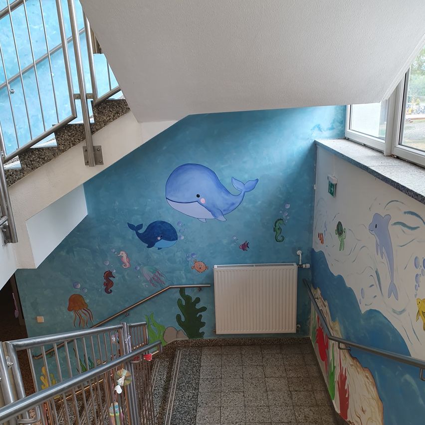 Ein Flur mit Treppen hat eine Wandmalerei mit einem Wal und Meerestieren an den Wänden. Es gibt einen weißen Heizkörper und Metallhandläufe auf beiden Seiten.