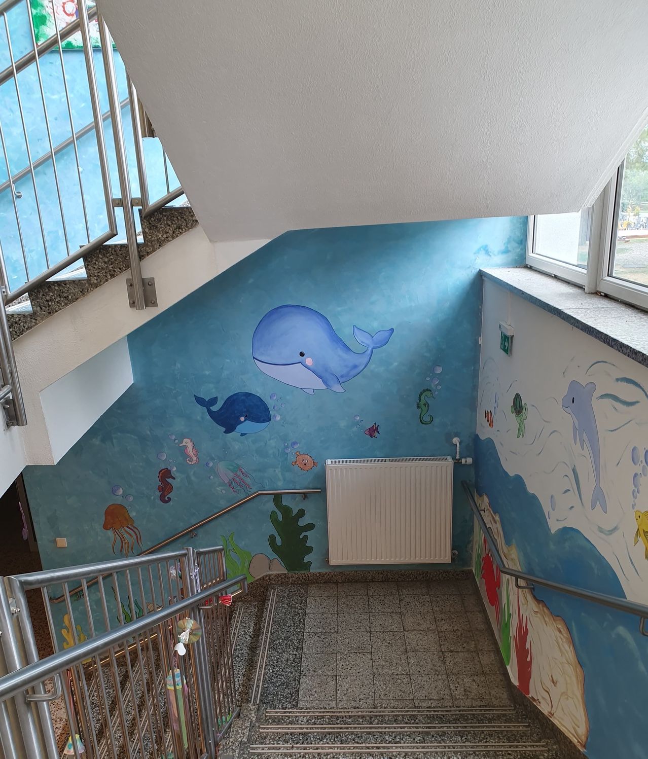Ein Flur mit Treppen hat eine Wandmalerei mit einem Wal und Meerestieren an den Wänden. Es gibt einen weißen Heizkörper und Metallhandläufe auf beiden Seiten.