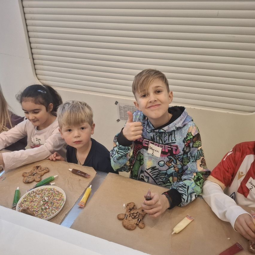Fünf Kinder sitzen um einen Tisch herum und sind in einer Backaktivität beschäftigt. Jedes Kind hat einen Keks vor sich, mit dekorativen Gegenständen wie Streuseln und Zuckerguss sichtbar. Ein Kind, das eine Kapuzenjacke trägt, gibt einen Daumen hoch. Hinter ihnen ist eine Wand mit Jalousien und einem Plakat zu sehen.