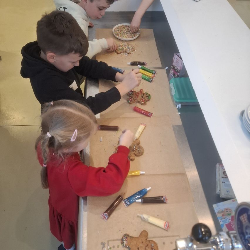 Kinder dekorieren Lebkuchen mit Zuckerguss und Streuseln auf einem Tisch. Ein Junge in einer schwarzen Kapuzenjacke und ein Mädchen in einem roten Kleid. Sie sind von verschiedenen Dekorationsmaterialien umgeben.
