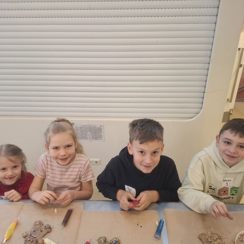 Vier Kinder sitzen an einem Tisch und basteln an Lebkuchenplätzchen. Jedes Kind hält Werkzeuge in der Hand und scheint konzentriert zu arbeiten. Hinter ihnen ist ein Fenster mit Jalousien und eine Tafel zu sehen.