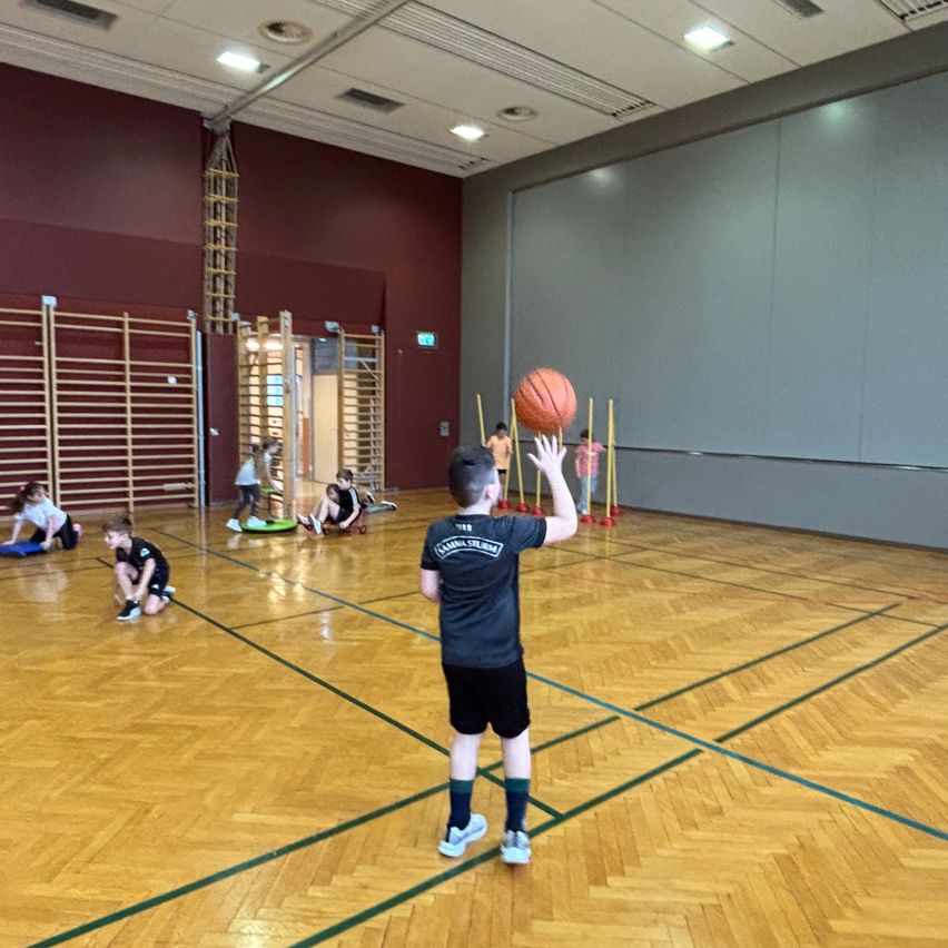 Ein Basketballspieler wirft einen Ball in einer Halle. Andere Spieler üben in der Nähe. Holzbarrieren und rote Stangen sind auf dem Spielfeld aufgestellt.