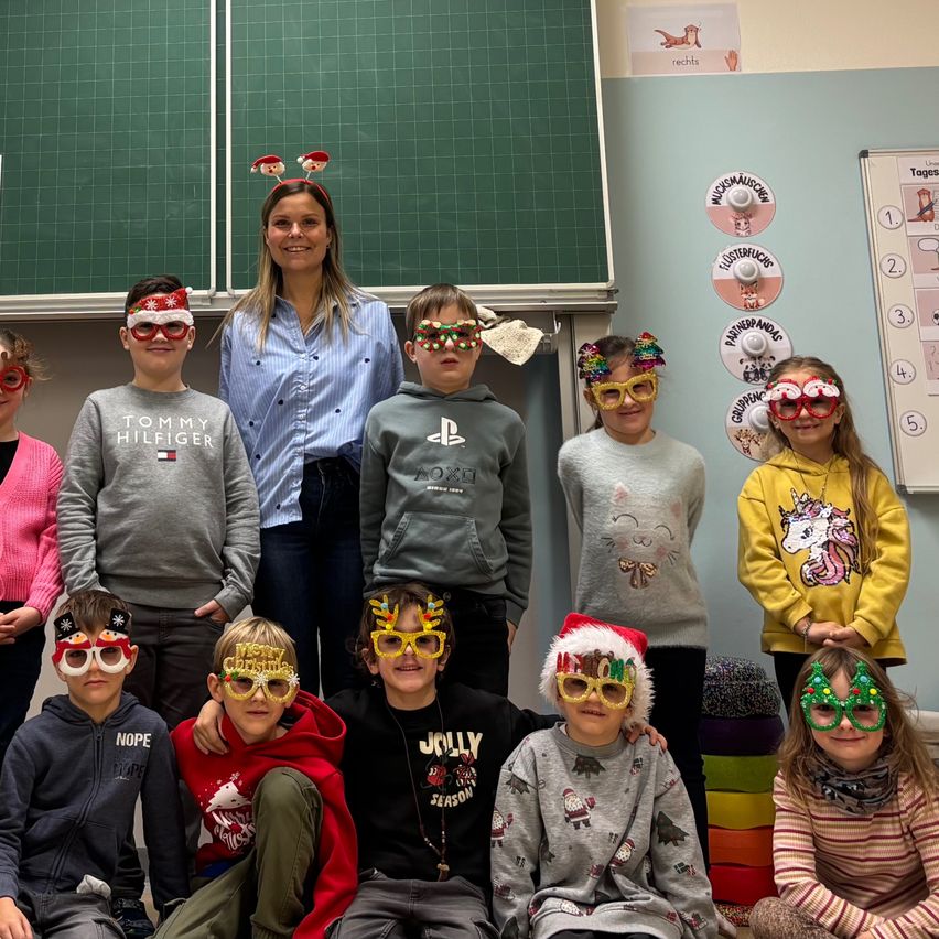 Eine Gruppe von Kindern und einer Frau posiert für ein Foto. Die Kinder tragen Gläser mit Weihnachtsdekorationen und die Frau lächelt. Hinter ihnen steht eine Tafel mit einigen Zeichnungen. Auf der rechten Seite befinden sich runde Aufkleber mit Bildern und Text.