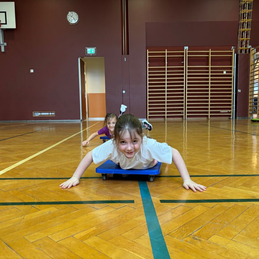 Zwei junge Mädchen sind in einer Turnhalle, ein Mädchen ist auf einem Schlitten, während das andere Mädchen hinter ihr ist. Sie lächeln.