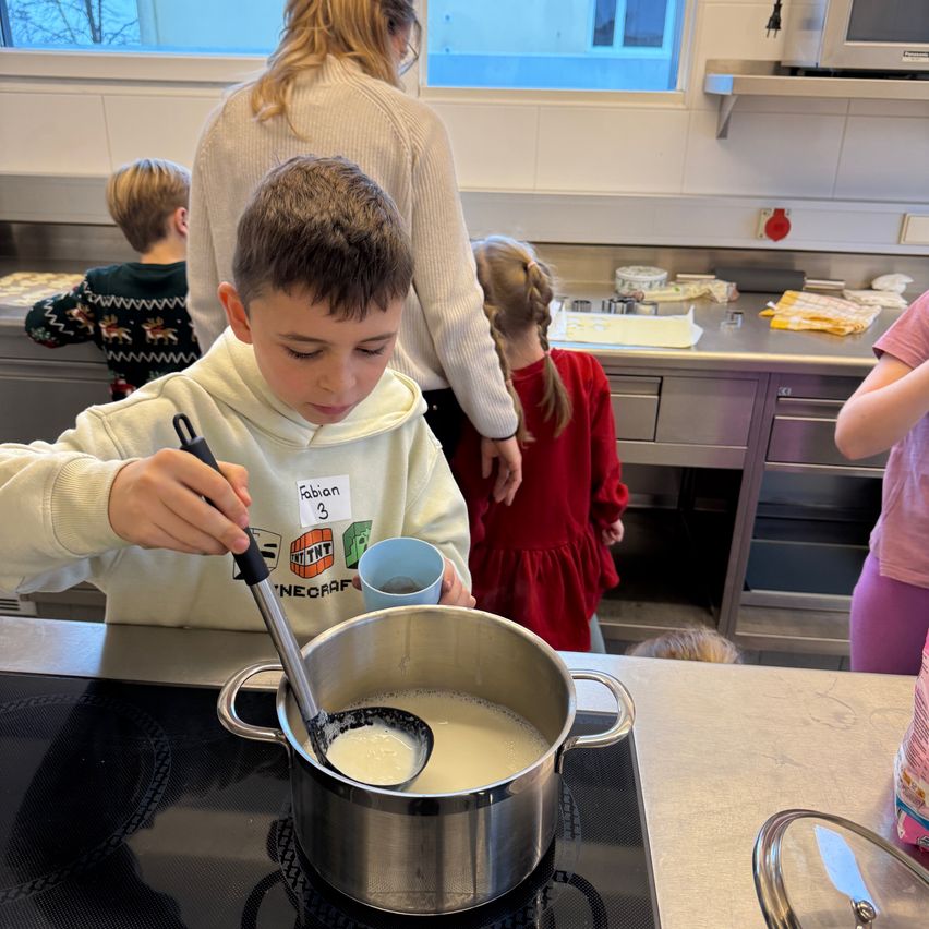 Ein Junge in einer weißen Hoodie mit dem Namen 'Fabian' rührt in einem Topf auf einem Herd, während eine Frau und zwei Kinder zusehen. Die Szene spielt sich in einer Küche ab.
