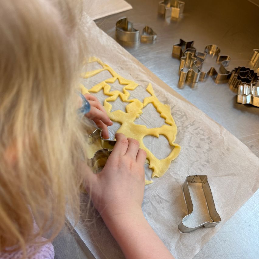 Ein kleines Mädchen benutzt Ausstecher, um stern- und herzförmige Kekse auf einem mit Papier bedeckten Tisch zu kreieren, mit weiteren Ausstechern um sie herum.